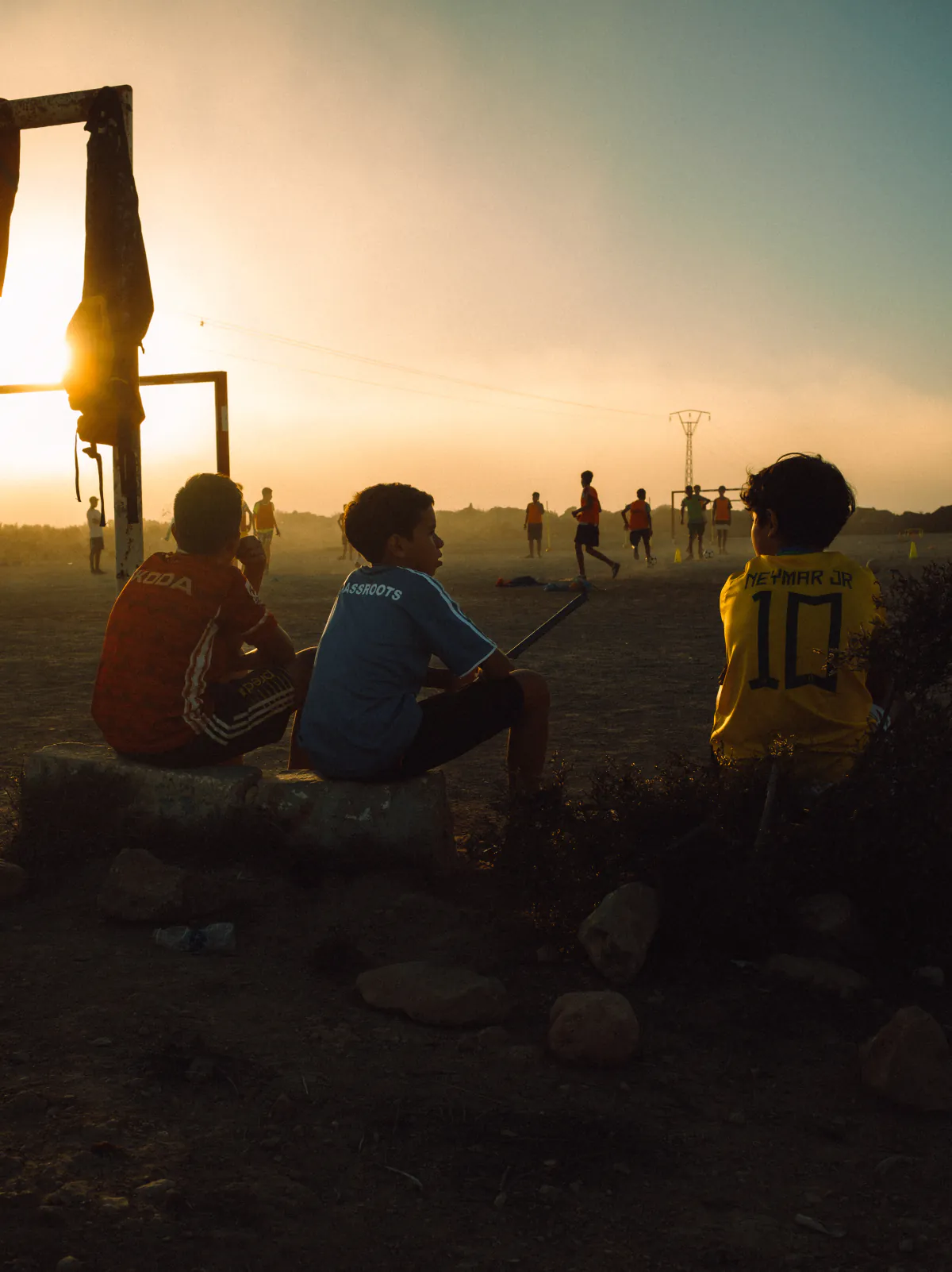 Kinder sitzen bei Sonnenuntergang am Rand eines staubigen Fußballplatzes