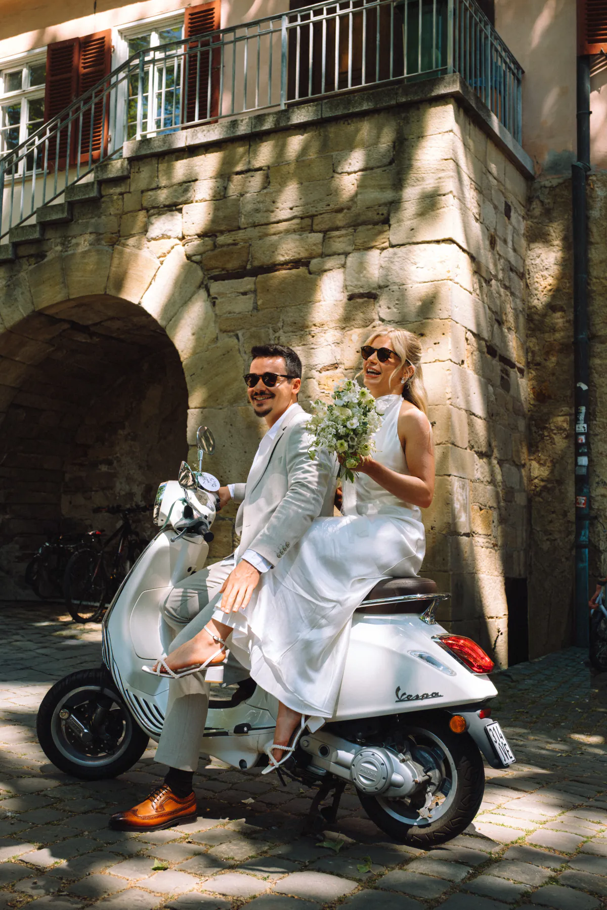 Brautpaar mit Sonnenbrillen auf einer weißen Vespa vor einem Steintor